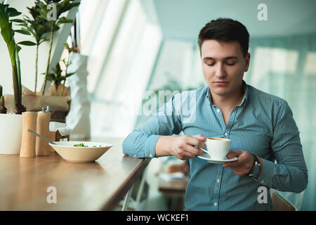 Beau jeune homme boire son café chaud et manger salade pour déjeuner pendant la pause au café près de l'office. Banque D'Images