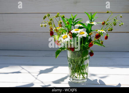 Branches de fraise mûre, rouge, blanc les marguerites et les feuilles de menthe dans un verre d'eau sur un moignon de bois Banque D'Images