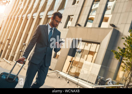 Toujours disponible. Jeune et beau bearded man in suit pulling suitcase et en regardant son smartphone pendant la marche à l'extérieur. Le voyage. Concept d'affaires Banque D'Images