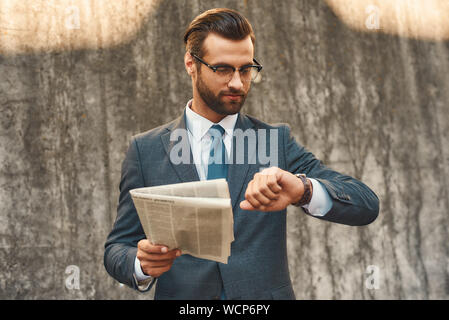 Quelle heure est-il jeune et beau homme barbu à lunettes contrôler le temps et la tenue de journal dans une main tout en se tenant contre un mur en pierre grise à l'extérieur. Concept d'entreprise. Concept de temps Banque D'Images