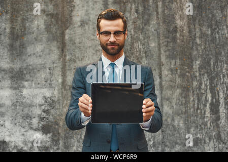 Homme d'affaires moderne. Portrait of handsome businessman barbu à lunettes montrant digital tablet at camera debout contre un mur en pierre grise à l'extérieur. Concept numérique. Concept d'affaires Banque D'Images