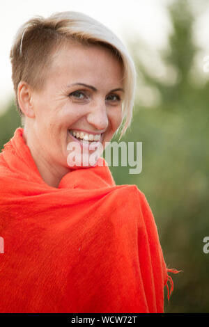 Close up of a middle-aged woman enveloppé dans une serviette orange après la baignade dans la rivière et souriant à la caméra, selective focus Banque D'Images