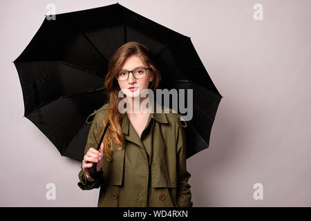 Une fille dans un manteau vert et les verres se dresse sur un fond blanc avec un parapluie noir et regarde la caméra. Banque D'Images