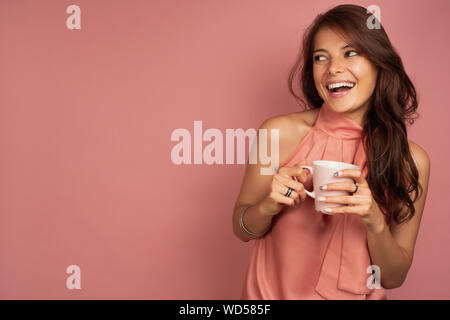 Une jeune fille brune dans une robe rose avec une tasse dans sa main se dresse sur un fond rose et semble rire joyeusement sur le côté. Banque D'Images