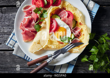 Crêpes avec œuf frit, jambon, tranches de tomates et de concombre frais dentaires sur une plaque blanche sur une vieille table en bois, vue de dessus, flatlay, close-up Banque D'Images