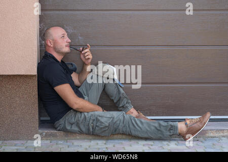 Un homme d'âge moyen repose sur le trottoir et le tabagisme. Portrait of caucasian homme chauve, à l'extérieur, Close up. De mauvaises habitudes, la toxicomanie. Li malsain Banque D'Images