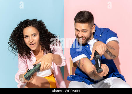 Jeune couple émotionnelle dans des vêtements décontractés posant sur fond rose et bleu. Concept d'émotions humaines, soins du visage, expession relations, ad. L'homme et la femme jouer jeux vidéo, essayez de gagner. Banque D'Images