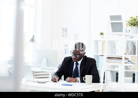African businessman in lunettes assis à la table et d'écrire quelque chose à l'office de tourisme Banque D'Images