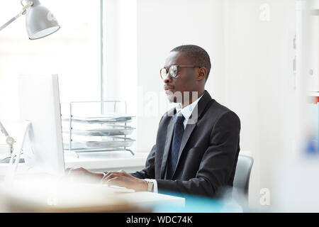 Gestionnaire de bureau Afrique de concentré à la table à la recherche à l'écran de l'ordinateur et en tapant tout en travaillant au bureau de la lumière Banque D'Images