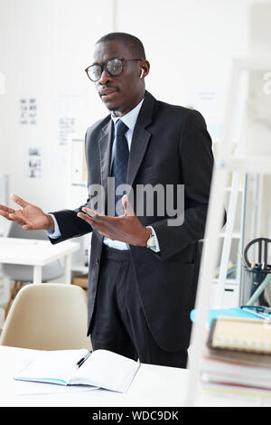 African Woman in suit, debout près de la table avec le bloc-notes portant un casque sans fil Bluetooth et parler dans at office Banque D'Images