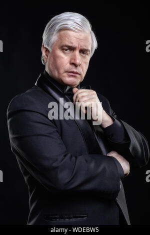 Studio portrait of senior man avec un visage sérieux expression sur fond noir. Homme mature en costume noir et chemise avec noeud papillon. À la recherche à Banque D'Images