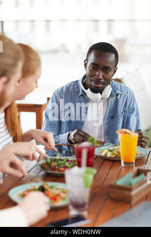 Jeune africain l'homme assis à la table en train de manger de la salade et de parler à ses amis qu'ils ont déjeuner au restaurant Banque D'Images