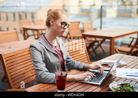 Red haired woman grave dans les lunettes travaillant comme pigiste elle assis et de la saisie sur ordinateur portable pendant le déjeuner d'affaires en outdoor cafe Banque D'Images