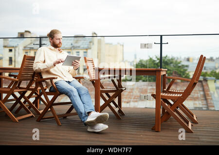 Jeune homme barbu assis sur une chaise et using digital tablet durant son temps libre à outdoor cafe Banque D'Images