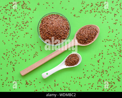 Cuillères en bois et céramique et bol en verre sont remplis de graines de lin entières sur un fond vert avec linseeds dispersés. La prévention des maladies du cœur de la nourriture. Banque D'Images