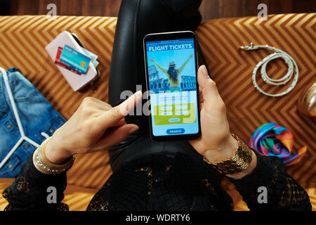Femme au foyer élégant assis sur un canapé dans le salon moderne avion réservation des billets en ligne sur un smartphone. Banque D'Images