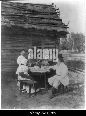 Groupe de paysans Russes posées à une table d'extérieur Résumé : Mise en scène genre vue inclut l'homme avec de la balalaïka, samovar sur la table, verser à boire, l'homme garçon par fenêtre et d'autres assis à table à l'extérieur d'une maison au toit de chaume. Banque D'Images