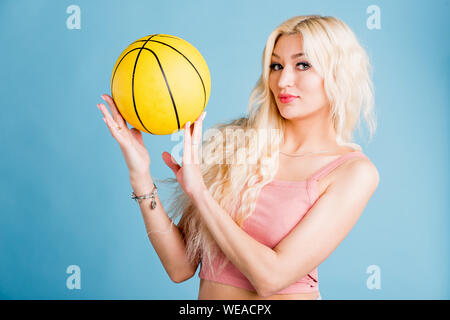 Jeune femme séduisante avec de longs cheveux blonds, beau maquillage, manucure portant haut court rose holding basketball looking at camera with pleasant expressi Banque D'Images