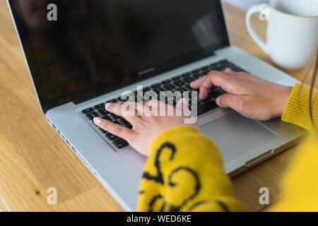 Vue du dessus de la main d'une femme de la saisie sur un ordinateur portable, sur un comptoir en bois en arrière-plan et un mug sur le côté. Banque D'Images