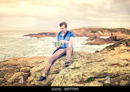 Homme assis en plein air sur la falaise de la lecture d'un e-book par la mer Banque D'Images
