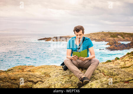 Homme assis en plein air sur la falaise de la lecture d'un e-book par la mer Banque D'Images