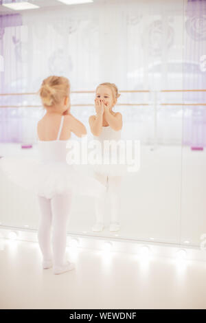 Petite fille ballerine en tutu blanc à côté du miroir avec son reflet dans elle à un studio de danse. Concept de ballet et d'enfants Banque D'Images