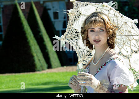 Belle femme Vêtements vintage assis sur la pelouse en face de stately home holding parasol et de boire du vin mousseux Banque D'Images