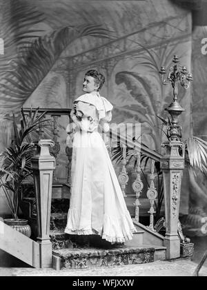 Un vintage style victorien ou au début de l'Edwardian Photographie noir et blanc montrant une jeune femme dans une belle robe à la mode de l'époque, posant dans un studio photographique, avec son dos légèrement pour l'appareil photo, comme elle regarde par dessus son épaule. Elle est debout sur un escalier. Banque D'Images