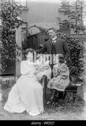 Un vintage style victorien ou au début de l'Edwardian Photographie noir et blanc montrant un groupe familial d'un père, mère, et deux filles, posant pour l'appareil photo à l'extérieur, dans un jardin de banlieue en Angleterre. Ils portent des vêtements typiques de l'époque. Banque D'Images