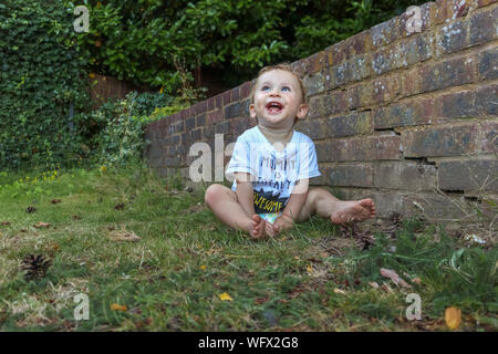 Une petite baby boy (de moins de 1 ans, white caucasian) bénéficiant en personne jouant dans le jardin assis par un mur de brique lève la tête et rit Banque D'Images