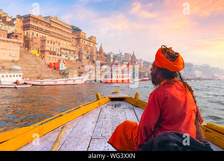 Sadhu baba assis sur un bateau en bois avec vue sur l'architecture historique de la ville de Varanasi avec Gange ghat au coucher du soleil. Banque D'Images