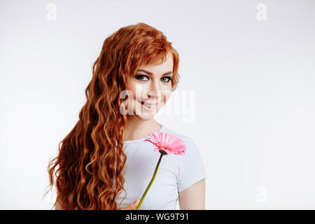 Closeup portrait of smiling, belle cute woman in casual vêtements avec sourire. de long cheveux bouclés, gingembre holding flower en mains près de joue, l'article o Banque D'Images