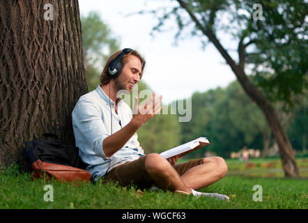 Cheerful student avec des écouteurs dans le parc du campus de lire un livre. Banque D'Images