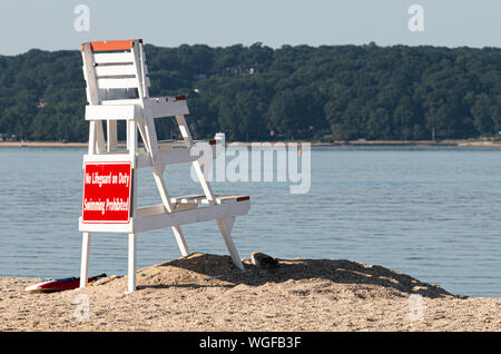 Un sauveteur blanc debout sur le sable à la recherche sur l'eau vide de la rive nord de Long Island, avec des collines et des arbres en arrière-plan. Banque D'Images