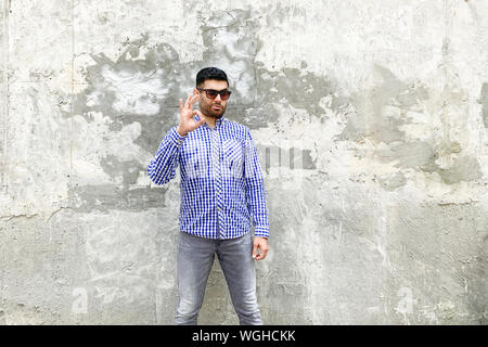 Son ok. Portrait de satisfait beau jeune homme barbu en chemise bleue à carreaux et des lunettes debout contre un mur gris béton, montrant un signe ok Banque D'Images