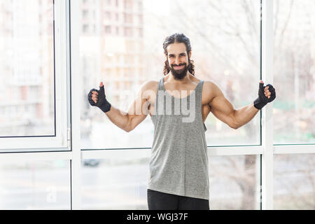 Portrait de jeunes adultes sportive positive satisfait formateur musculaire homme avec de longs cheveux bouclés avec commandes d'armes lourdes, showing Thumbs up et smi Banque D'Images