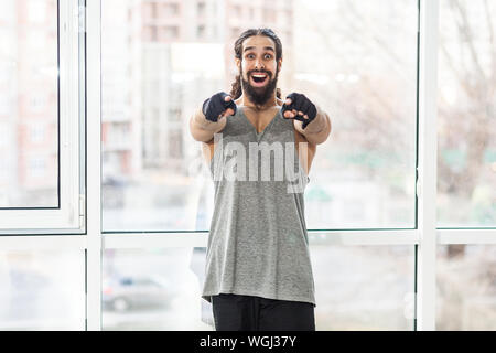 Portrait de jeunes adultes heureux surpris formateur musculaire homme avec les cheveux longs bouclés, permanent pointer du doigt à vous avec de grands yeux, looking at camera wifi Banque D'Images