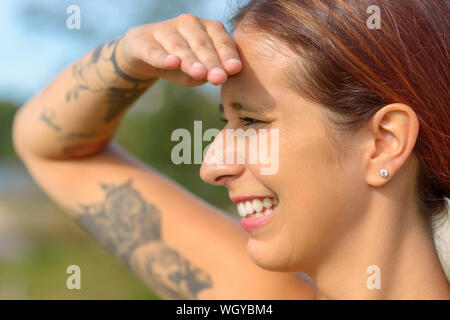 Jolie femme gardant un oeil dehors avec sa main posée sur son front pour protéger ses yeux du soleil et un sourire heureux Banque D'Images