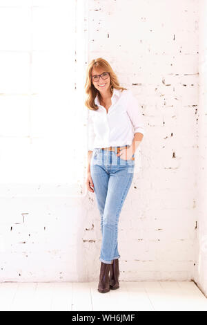 Shot pleine longueur à maturité attrayante femme portant une chemise blanche et un jean bleu tout en se tenant à mur blanc. Banque D'Images