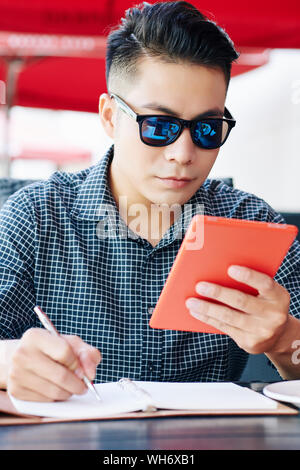 Jeune homme sérieux dans les lunettes de lecture d'informations sur l'ordinateur tablette et prendre des notes dans planner Banque D'Images