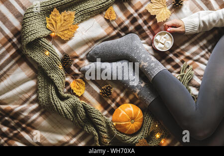Les mains et les pieds de femmes chandail en laine et chaussettes gris confortable holding tasse de café chaud avec de la guimauve, assis sur la citrouille avec plaid, en écharpe, Banque D'Images