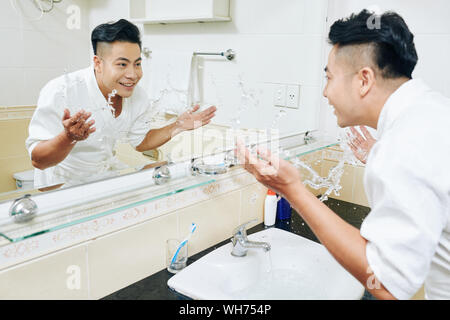 Les jeunes Vietnamiens positive heureux homme en peignoir aux projections d'eau de couler sur son visage pour se réveiller du sommeil formulaire Banque D'Images