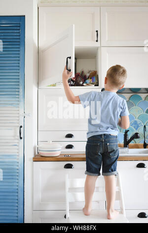 Vue arrière de petit garçon debout sur une chaise dans la cuisine à la placard dans Banque D'Images