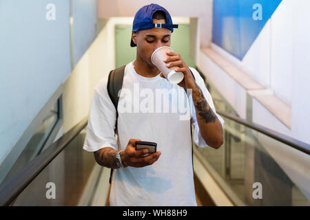 Jeune homme tatoué avec sac à dos, debout sur l'escalator à la recherche de téléphone cellulaire pour aller boire du café Banque D'Images