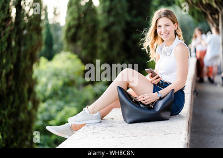 Portrait of smiling young woman with headphones et smartphone assis sur un mur Banque D'Images