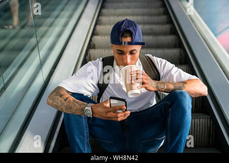 Jeune homme avec sac à dos, assis sur l'escalator à la recherche de téléphone cellulaire pour aller boire du café Banque D'Images