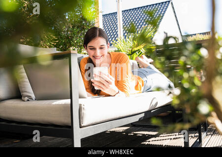 Young woman lying on couch sur terrasse à l'aide d'un téléphone cellulaire et ordinateur portable Banque D'Images