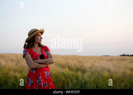 Woman wearing Straw Hat et Red robe d'été avec floral design standing in front of grain field Banque D'Images