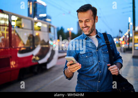 Man using smartphone et écouter de la musique tout en se tenant à l'arrêt dans la soirée Banque D'Images