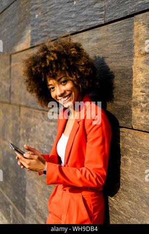 Portrait of happy young woman with smartphone portant à la mode tailleur-pantalon rouge leaning against wall Banque D'Images